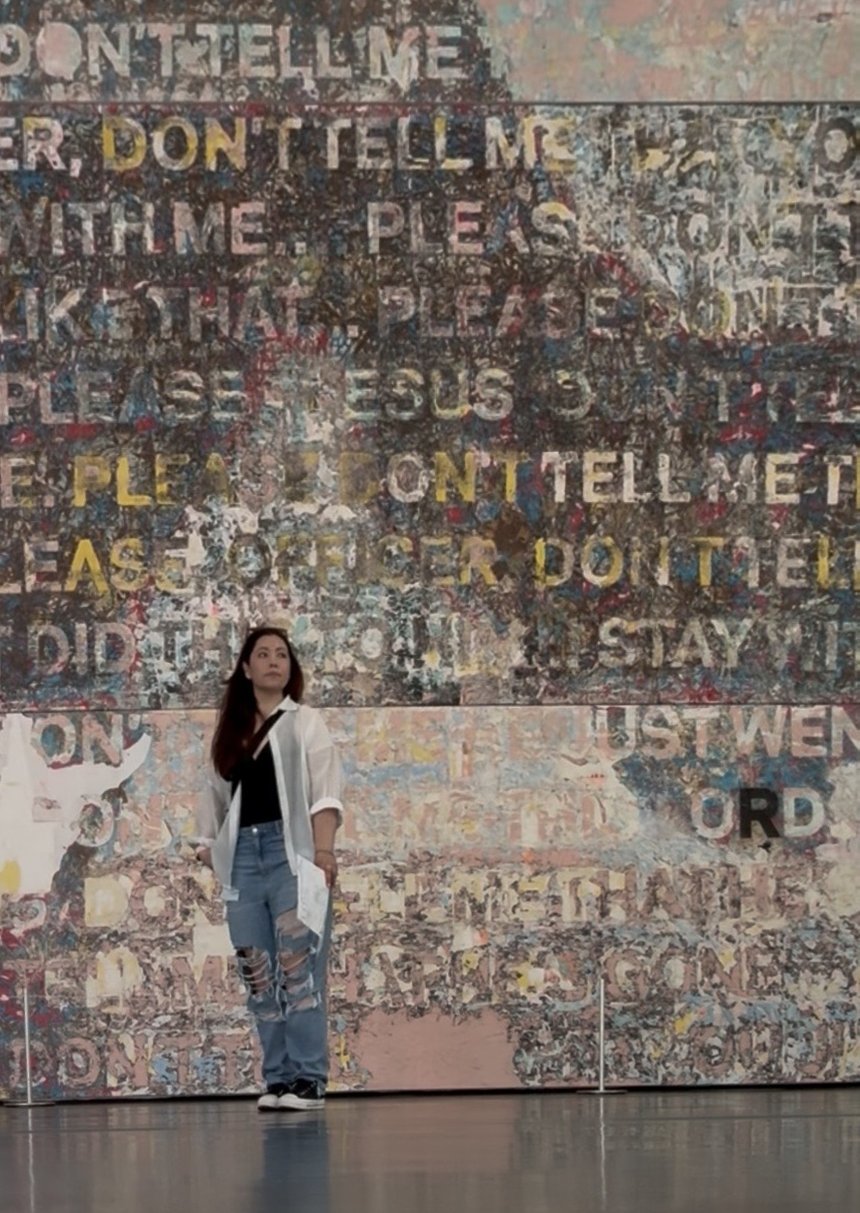 Karen Ballesteros standing in front of a large mixed-media typographic mural at a gallery